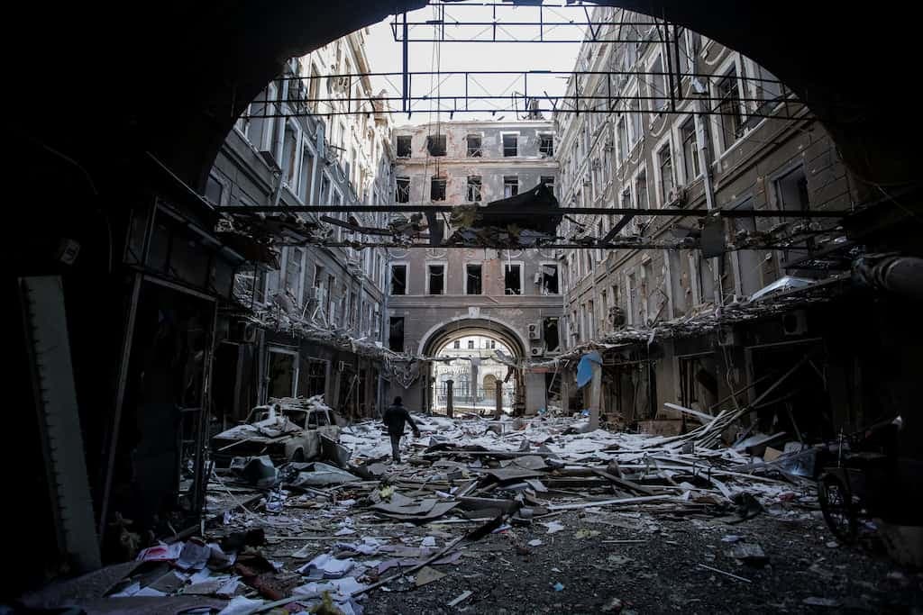 Разрушенный внутренний двор или проход между зданиями. Камера смотрит через тёмную арку наружу, где на земле лежит гора обломков и строительного мусора. Фасады зданий вокруг сильно повреждены, многие окна разбиты. В центре руин видна фигура идущего человека.