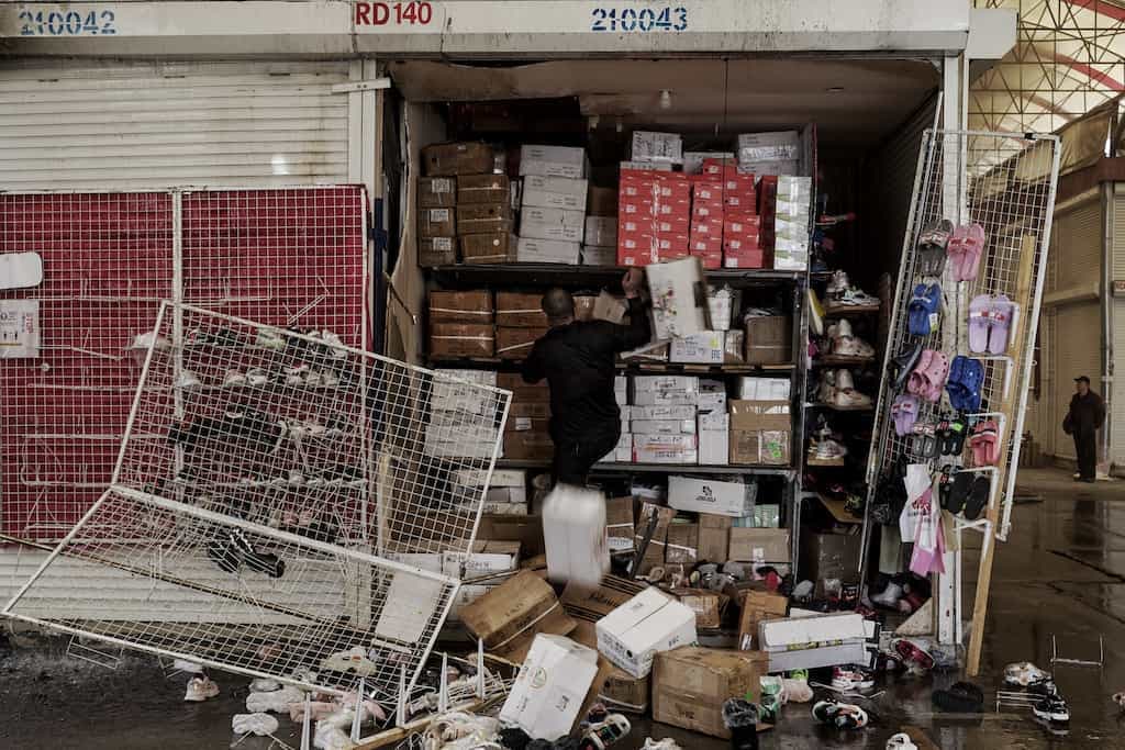 Мужчина в тёмной одежде стоит внутри открытого складского помещения или торгового павильона, заполненного коробками, обувью и другими товарами, сложенными на полках. Перед павильоном на мокром полу разбросаны товары и опрокинутая металлическая решётка-стенд. На заднем плане справа видна ещё одна фигура.