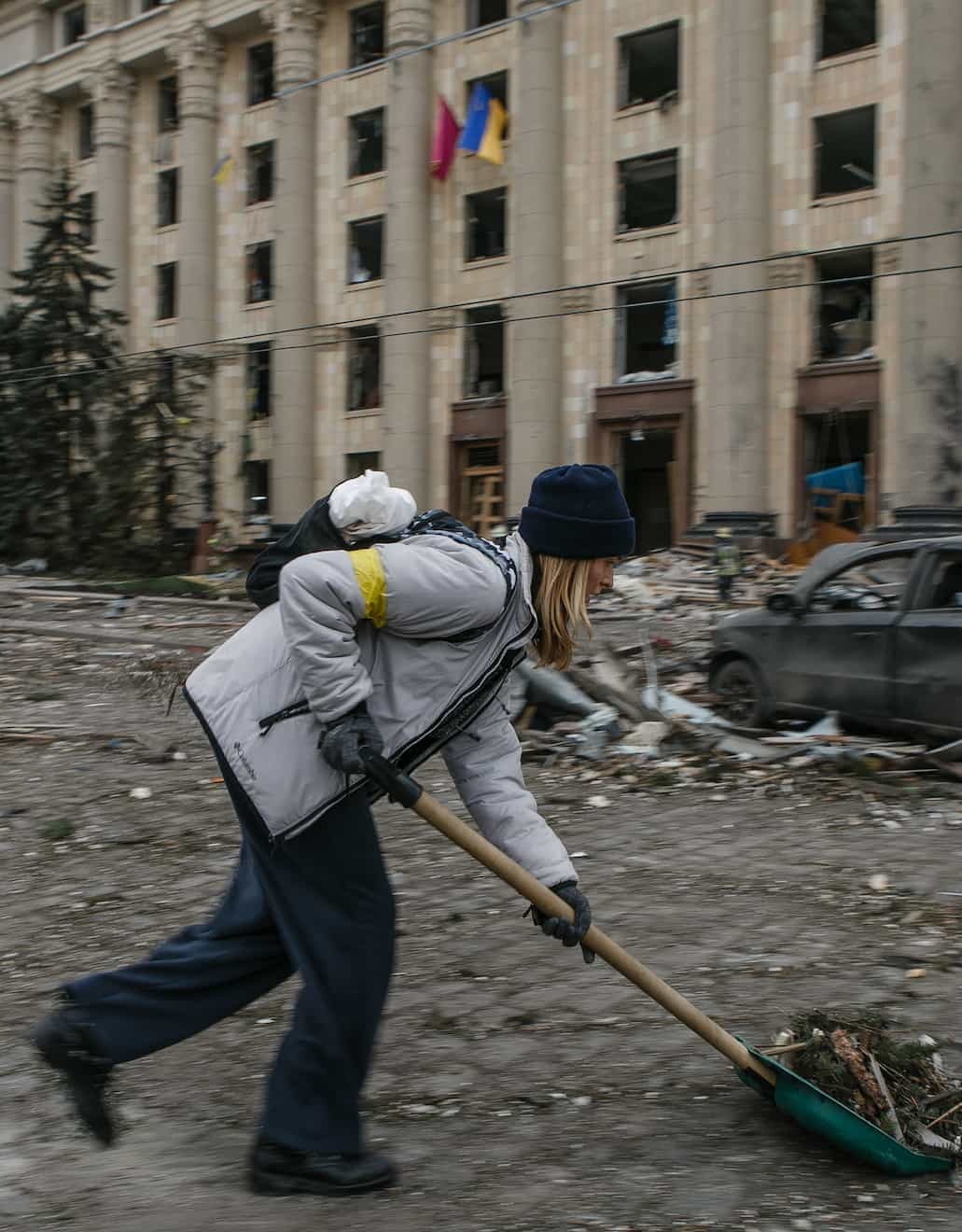 Женщина убирает мусор лопатой на фоне здания с украинским флагом.